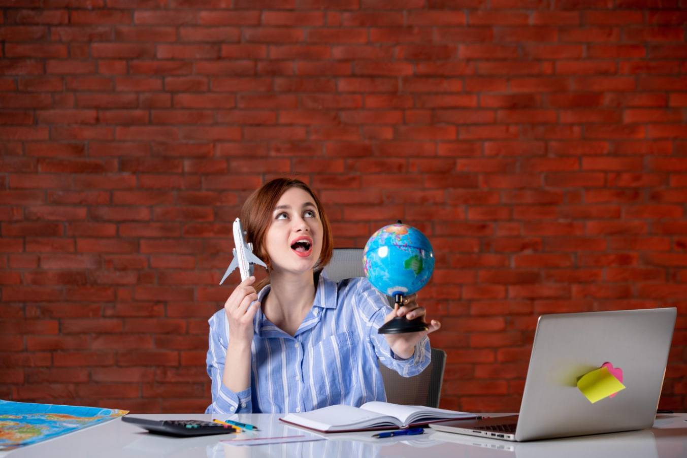 A woman sits at a desk in front of a red brick wall, playfully holding a toy airplane and a small globe while looking upward with an excited expression